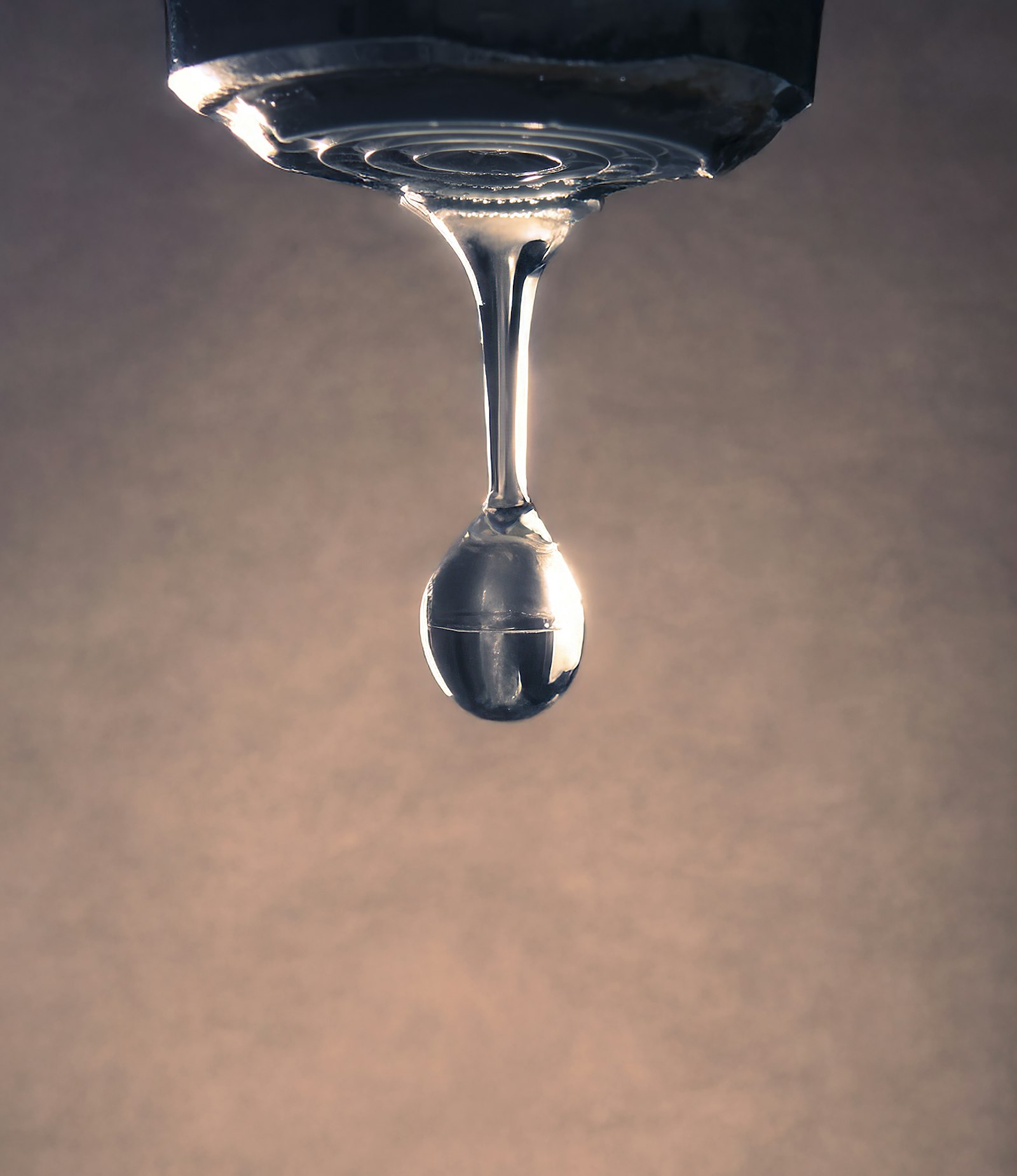 Primer plano de una gota de agua colgando de un grifo metálico con luz cálida de fondo